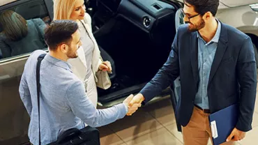 Three people near a car, two shaking hands.