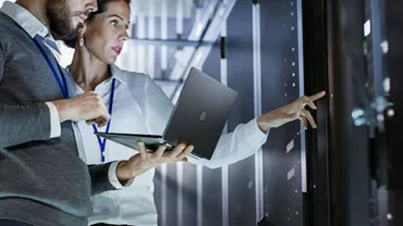 Two people in a server room reviewing information on a laptop.