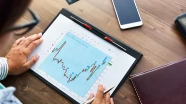 Person analyzing stock chart on tablet next to notebook and phone.
