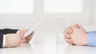 Two people sit across a table, one holding a paper.