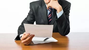 Person in a suit reads a document at a wooden table.