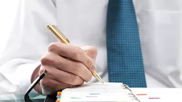 Person writing in a planner with a pen, wearing a blue tie.