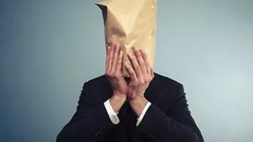 Person in a suit with a paper bag over their head, standing against a plain background.