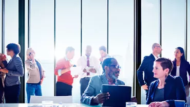 People in a bright office engaged in discussions near large windows.