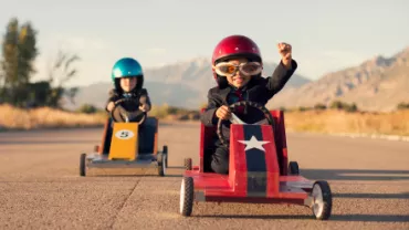 Children racing in toy cars with helmets, one cheering on a sunny day.