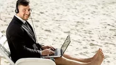 Person in a suit with headphones using a laptop on the beach.