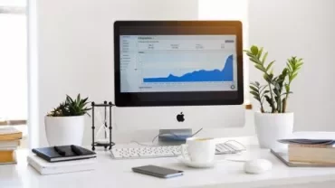A computer on a desk with a graph displayed on the screen, surrounded by plants and books.
