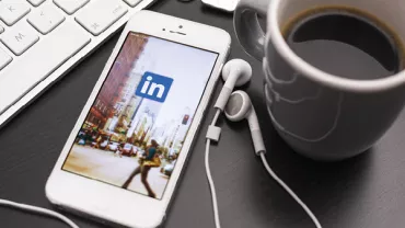 Smartphone with LinkedIn app next to a keyboard and coffee cup.