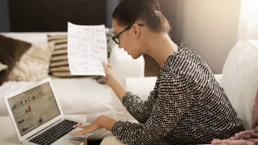 Person working on a laptop with a spreadsheet in hand.