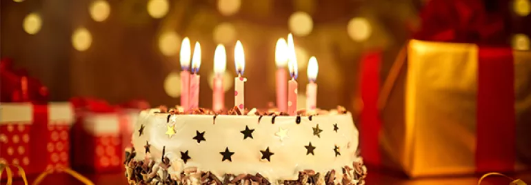 A birthday cake with lit candles and gift boxes in the background.