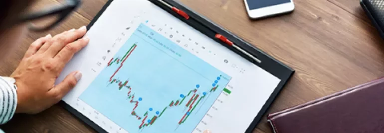 Personne analysant un graphique boursier sur une tablette à côté d'un carnet et d'un téléphone.