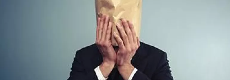 Personne en costume avec un sac en papier sur la tête, devant un fond uni.