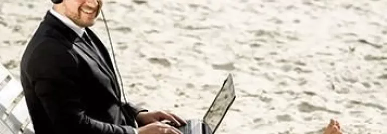 Person in a suit with headphones using a laptop on the beach.
