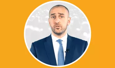 Person in a suit speaking with a microphone in front of a blurred background.
