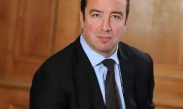 Person in a suit seated against a wooden paneled background.