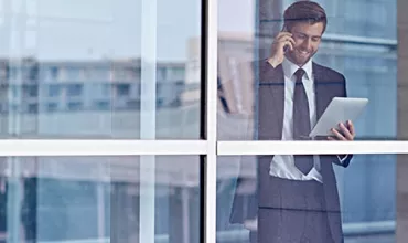 Person in office attire using a tablet and talking on phone by a window.